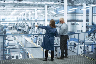 Two professionals observe a large industrial manufacturing facility from an elevated platform with expansive windows.