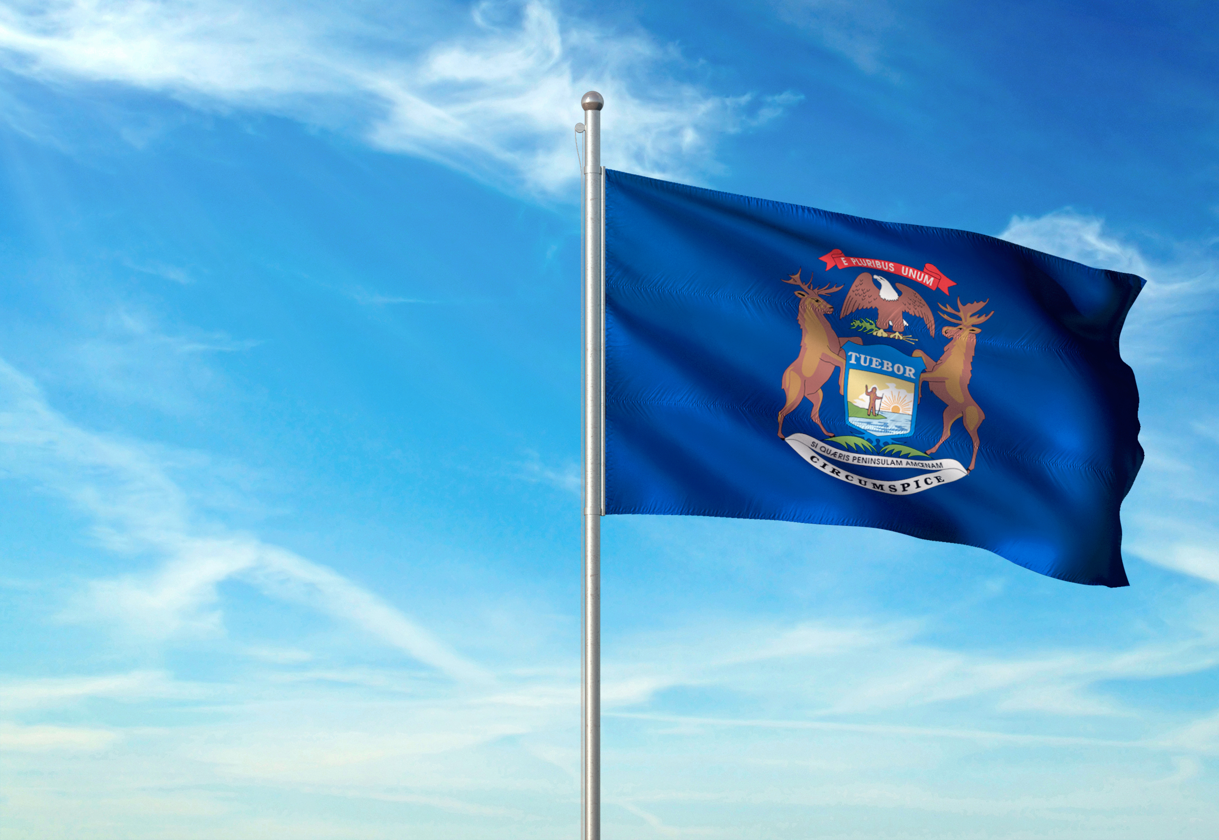 Michigan state flag waving on a flagpole against a blue sky with white clouds