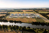 Aerial view of industrial facility with blue-roofed buildings, storage tanks, and surrounding agricultural fields near a river