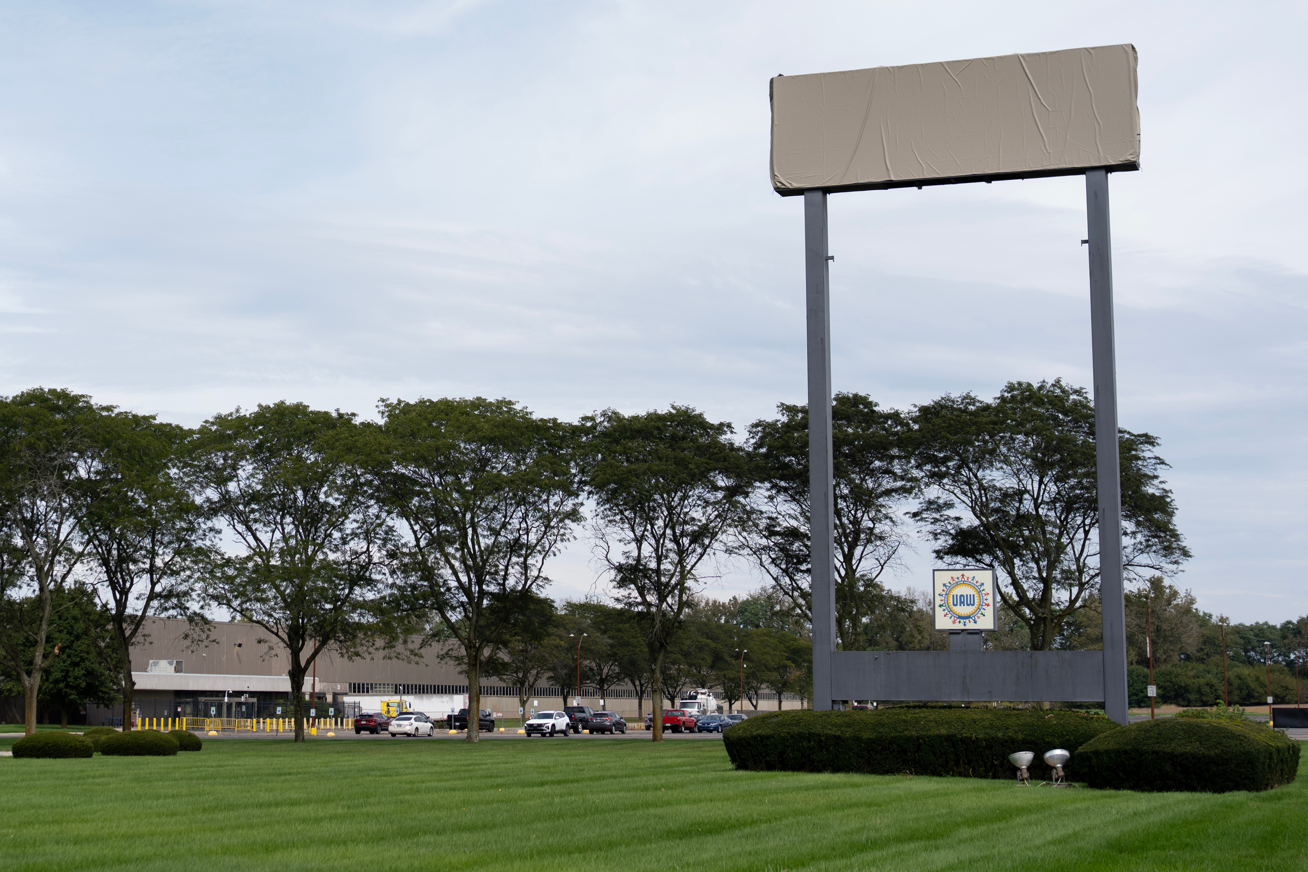 Large blank billboard with metal frame in commercial area surrounded by trees, buildings, and parked vehicles