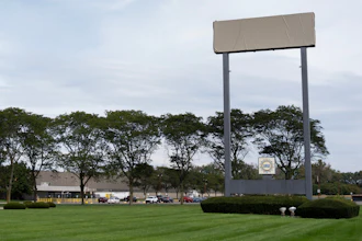 Large blank billboard with metal frame in commercial area surrounded by trees, buildings, and parked vehicles