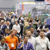 Crowded trade show floor with attendees wearing lanyards and ID badges networking among exhibition booths