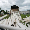 White PVC pipes being unloaded from a dump truck at an industrial yard with green buildings in background