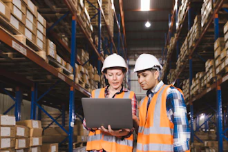 Two warehouse workers in safety gear reviewing inventory data on laptop in storage facility