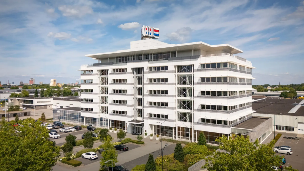 Modern white multi-story commercial building with glass windows and flags on rooftop, surrounded by landscaping and parked vehicles